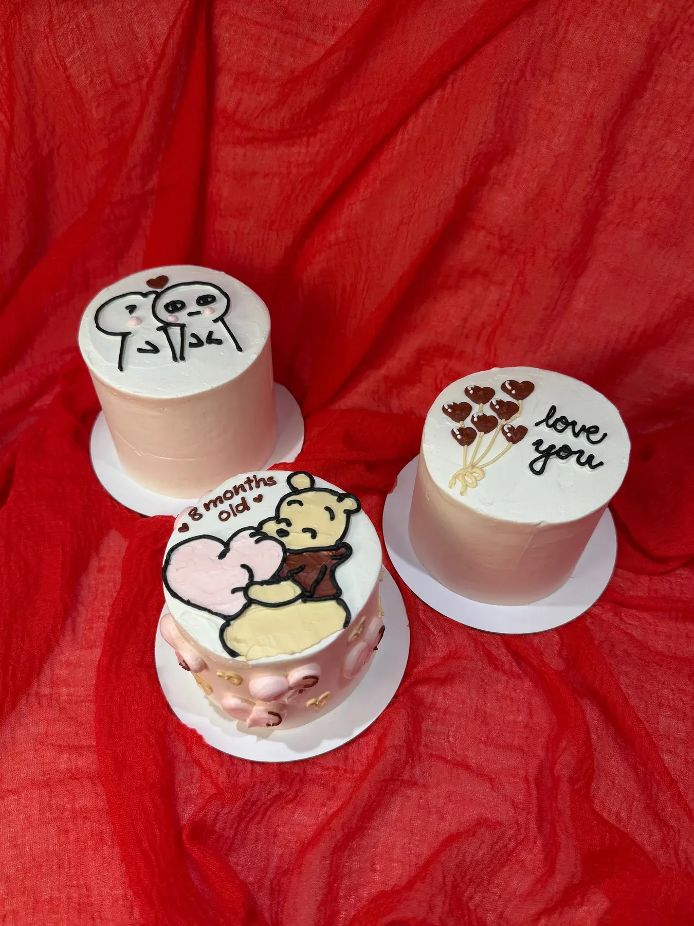 Three little bento cakes on red fabric, piped with a cartoon couple, a Winnie the Pooh “8 months old” and a heart-balloon “love you”