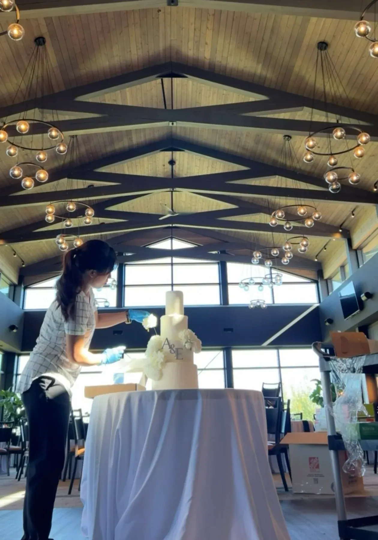 Nicole styling white flowers on a four-tier A and E wedding cake in a reception hall with a vaulted wooden ceiling