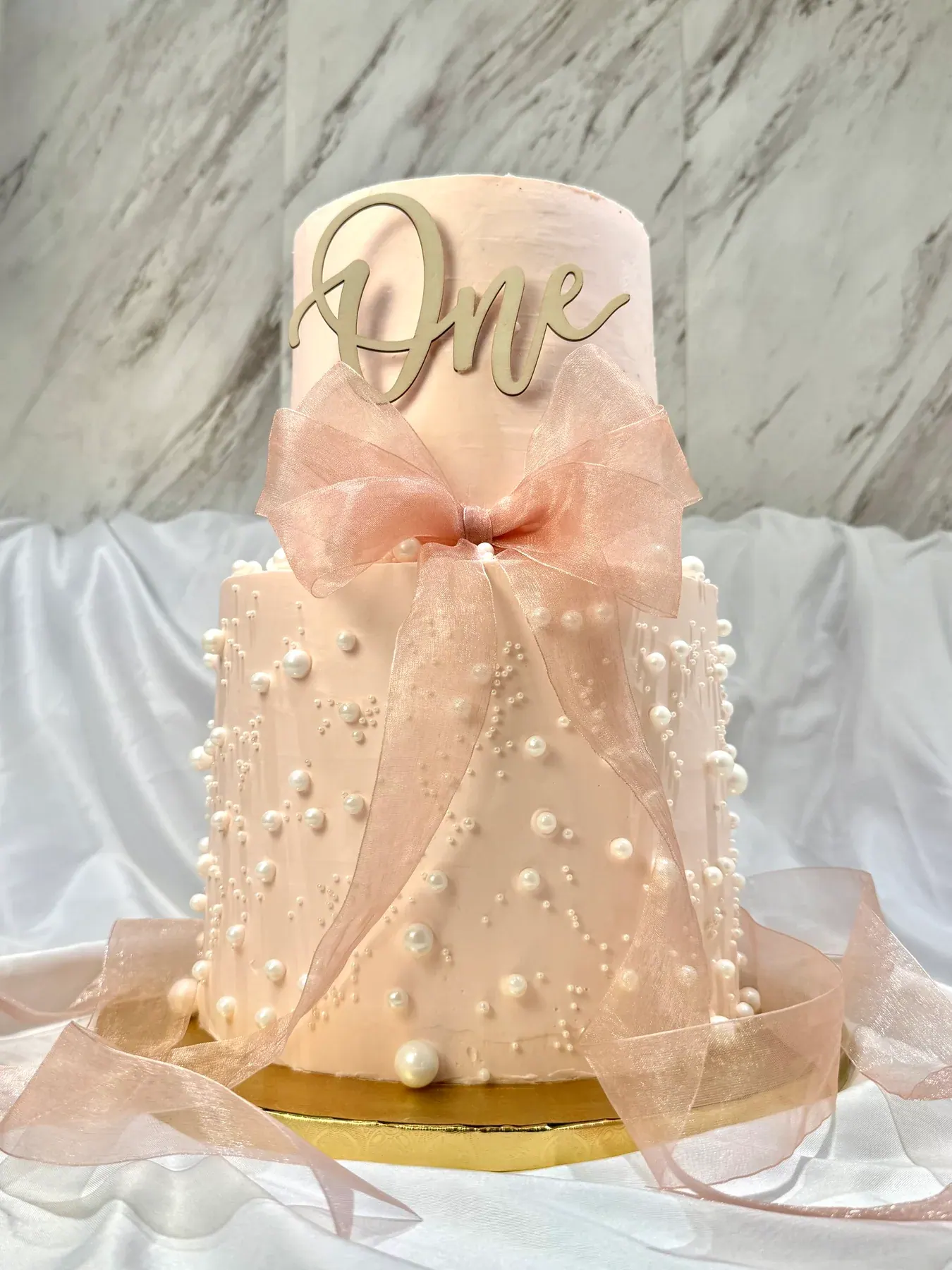 Nicole's blush two-tier birthday cake, scattered with pearls and tied with a sheer organza bow under a gold “One” topper