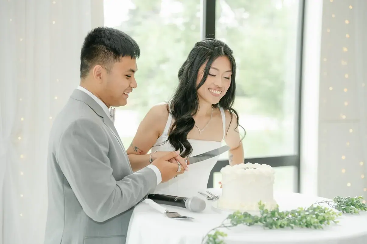 A bride and groom laughing as they cut into their white Nicsdelite wedding cake by a bright window